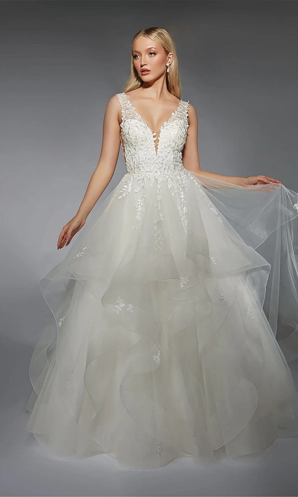 A woman in the Alyce 7126 long white dress in ivory, featuring a layered skirt, deep V-neckline, and lace details, stands against a gray background holding part of her tulle skirt to the side.